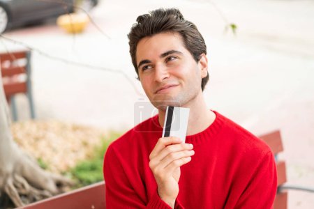 Foto de Joven hombre guapo sosteniendo una tarjeta de crédito y pensando - Imagen libre de derechos