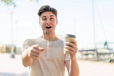 Téléchargez les photos : Jeune homme beau tenant un café à emporter à l'extérieur surpris et pointant devant - en image libre de droit