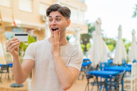 Photo for Young handsome man holding a credit card at outdoors with surprise and shocked facial expression - Royalty Free Image