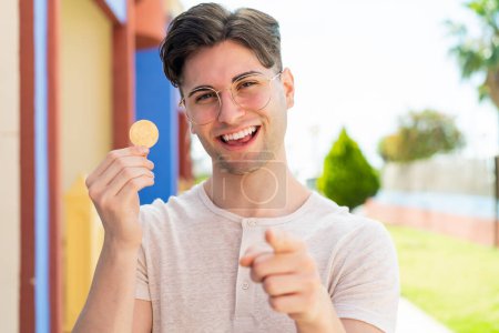 Foto de Joven hombre guapo sosteniendo un Bitcoin al aire libre apunta con el dedo a usted con una expresión de confianza - Imagen libre de derechos