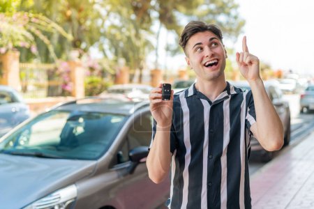 Téléchargez les photos : Jeune homme beau tenant les clés de voiture à l'extérieur dans l'intention de réaliser la solution tout en levant le doigt - en image libre de droit