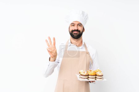 Téléchargez les photos : Jeune homme tenant gâteau muffin sur fond blanc isolé heureux et comptant trois avec les doigts - en image libre de droit