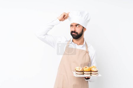 Téléchargez les photos : Jeune homme tenant gâteau muffin sur fond blanc isolé ayant des doutes tout en grattant la tête - en image libre de droit