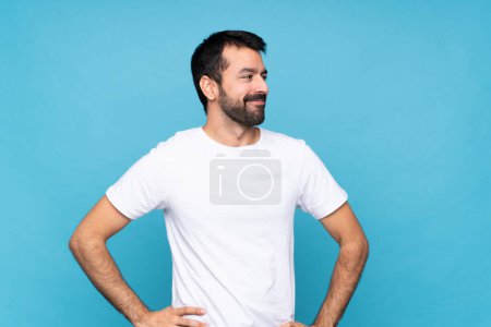 Foto de Joven con barba sobre fondo azul aislado posando con los brazos en la cadera y sonriendo - Imagen libre de derechos