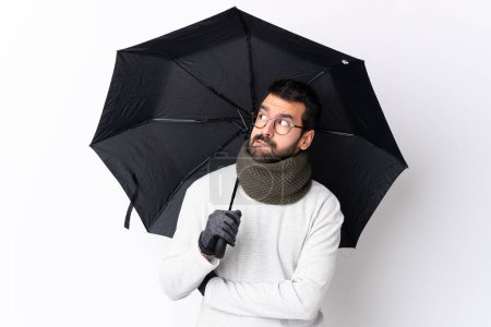 Photo for Caucasian handsome man with beard holding an umbrella over isolated white wall with confuse face expression - Royalty Free Image