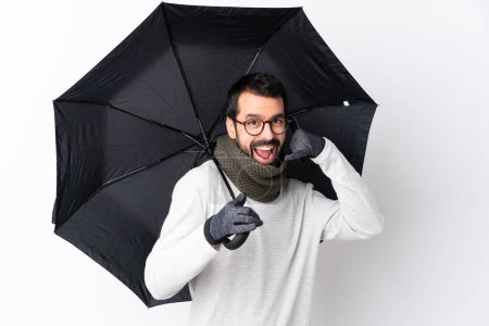 Foto de Hombre guapo caucásico con barba sosteniendo un paraguas sobre una pared blanca aislada haciendo un gesto telefónico y apuntando hacia delante - Imagen libre de derechos