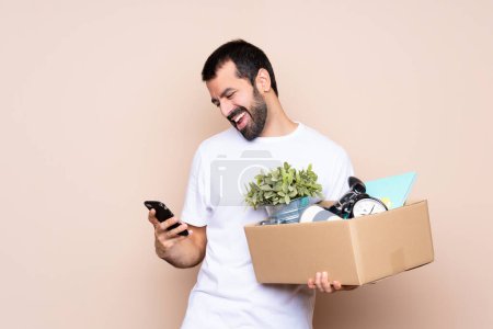 Foto de Hombre sosteniendo una caja y moviéndose en un nuevo hogar sobre un fondo aislado con teléfono en posición de victoria - Imagen libre de derechos