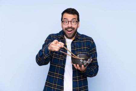 Photo for Young caucasian man over blue background surprised and pointing front while holding a bowl of noodles with chopsticks - Royalty Free Image