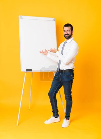 Photo for Full-length shot of businessman giving a presentation on white board over isolated yellow background extending hands to the side for inviting to come - Royalty Free Image