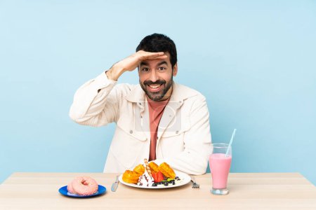 Photo for Man at a table having breakfast waffles and a milkshake looking far away with hand to look something - Royalty Free Image