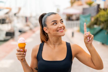 Téléchargez les photos : Jeune jolie femme avec une crème glacée cornet à l'extérieur dans l'intention de réaliser la solution tout en levant le doigt - en image libre de droit