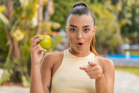 Téléchargez les photos : Jeune jolie femme avec une pomme à l'extérieur surpris et pointant devant - en image libre de droit