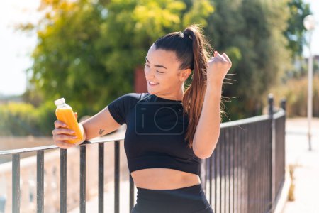 Photo for Young pretty sport woman holding an orange juice at outdoors celebrating a victory - Royalty Free Image