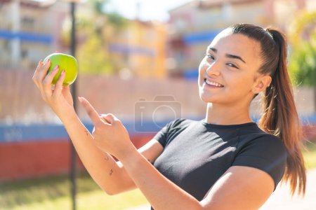 Téléchargez les photos : Jeune jolie femme brune avec une pomme à l'extérieur et le pointant - en image libre de droit