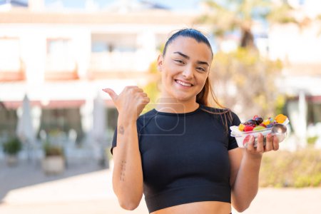 Téléchargez les photos : Jeune jolie femme brune tenant un bol de fruits à l'extérieur pointant vers le côté pour présenter un produit - en image libre de droit