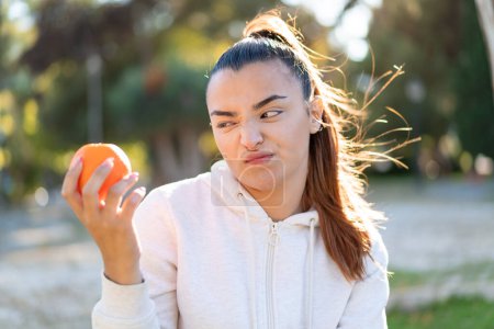 Téléchargez les photos : Jeune jolie femme brune tenant une orange à l'extérieur avec une expression triste - en image libre de droit