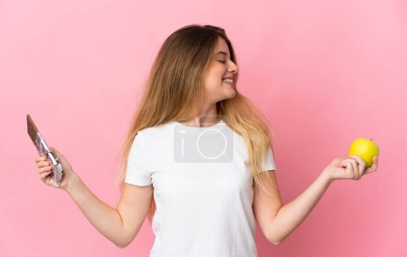 Téléchargez les photos : Jeune femme blonde isolée sur fond rose prenant un comprimé de chocolat dans une main et une pomme dans l'autre - en image libre de droit
