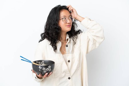 Foto de Joven mujer asiática aislada sobre fondo blanco que tiene dudas y con expresión de cara confusa mientras sostiene un tazón de fideos con palillos - Imagen libre de derechos