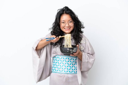 Téléchargez les photos : Jeune femme asiatique portant kimono isolé sur fond blanc tenant un bol de nouilles avec des baguettes - en image libre de droit