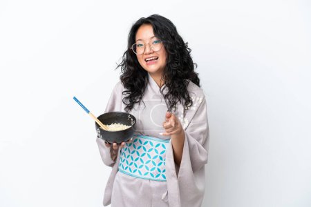 Téléchargez les photos : Jeune femme asiatique portant kimono isolé sur fond blanc pointe doigt à vous avec une expression confiante tout en tenant un bol de nouilles avec des baguettes - en image libre de droit
