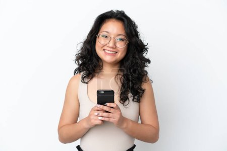 Foto de Joven mujer asiática aislada sobre fondo blanco mirando a la cámara y sonriendo mientras usa el móvil - Imagen libre de derechos