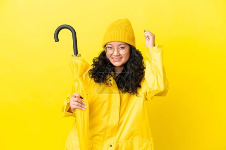 Asiatin mit regenfestem Mantel und Regenschirm auf gelbem Hintergrund feiert einen Sieg
