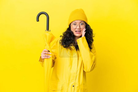 Asiatin mit regendichtem Mantel und Regenschirm auf gelbem Hintergrund mit Überraschung und schockiertem Gesichtsausdruck