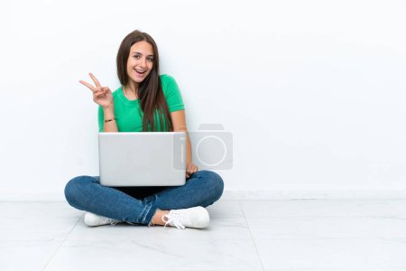 Téléchargez les photos : Jeune femme ukrainienne avec un ordinateur portable assis sur le sol souriant et montrant signe de victoire - en image libre de droit