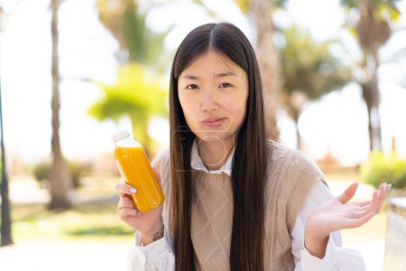 Téléchargez les photos : Jolie Chinoise tenant un jus d'orange à l'extérieur faisant des doutes geste tout en soulevant les épaules - en image libre de droit