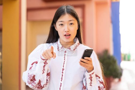 Téléchargez les photos : Jeune femme chinoise utilisant un téléphone mobile à l'extérieur surpris et pointant devant - en image libre de droit