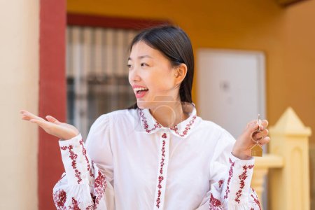 Téléchargez les photos : Jeune femme chinoise tenant les clés de la maison à l'extérieur avec expression faciale surprise - en image libre de droit