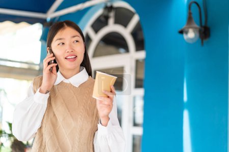 Téléchargez les photos : Jeune femme chinoise à l'extérieur en utilisant un téléphone mobile et tenant un café avec une expression heureuse - en image libre de droit