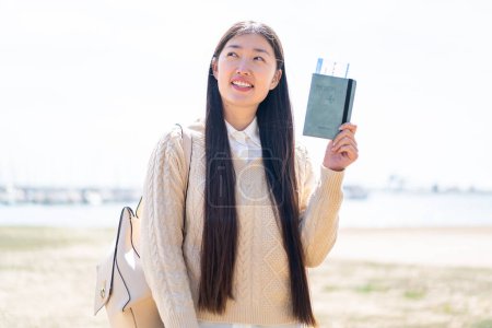 Téléchargez les photos : Jeune femme chinoise tenant un passeport à l'extérieur levant les yeux tout en souriant - en image libre de droit