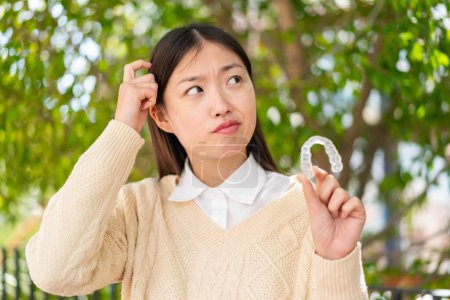 Téléchargez les photos : Jeune femme chinoise tenant des bretelles invisibles à l'extérieur ayant des doutes et avec une expression du visage confuse - en image libre de droit