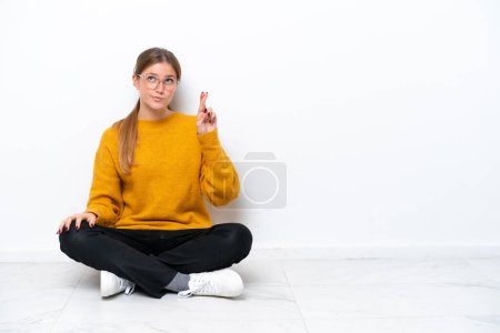 Téléchargez les photos : Jeune femme caucasienne assise sur le sol isolé sur fond blanc avec les doigts croisés et souhaitant le meilleur - en image libre de droit