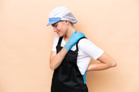 Fishmonger caucasian woman wearing an apron and holding a raw fish isolated on beige background suffering from pain in shoulder for having made an effort