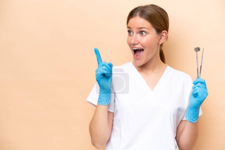Foto de Dentista mujer caucásica sosteniendo herramientas aisladas sobre fondo beige con la intención de realizar la solución mientras levanta un dedo - Imagen libre de derechos
