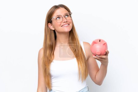 Photo for Young pretty blonde woman holding piggy bank isolated on white background looking up while smiling - Royalty Free Image
