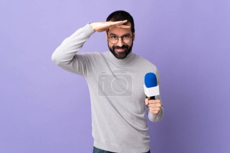 Foto de Hombre reportero adulto con barba sosteniendo un micrófono sobre un fondo púrpura aislado mirando lejos con la mano para mirar algo - Imagen libre de derechos