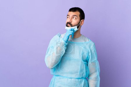 Foto de Cirujano con barba con uniforme azul sobre fondo púrpura aislado que tiene dudas y con expresión de cara confusa - Imagen libre de derechos