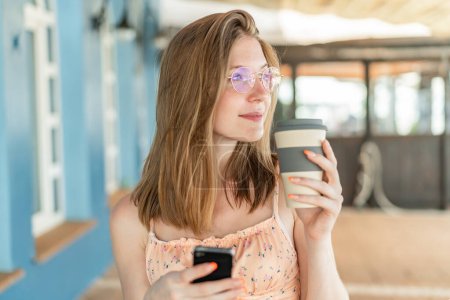 Junge Französin mit Brille im Freien mit Handy und Kaffee in der Hand