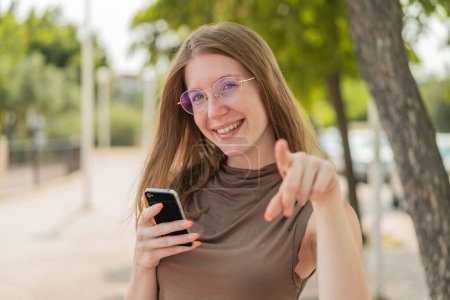 Téléchargez les photos : Française avec des lunettes utilisant un téléphone mobile à l'extérieur pointe du doigt vers vous avec une expression confiante - en image libre de droit