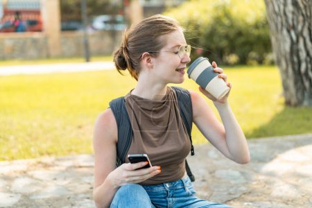 Foto de Joven chica francesa con gafas al aire libre utilizando el teléfono móvil y sosteniendo un café para llevar - Imagen libre de derechos
