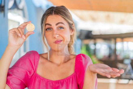 Téléchargez les photos : Jeune femme blonde tenant un Bitcoin à l'extérieur faisant des doutes geste tout en soulevant les épaules - en image libre de droit