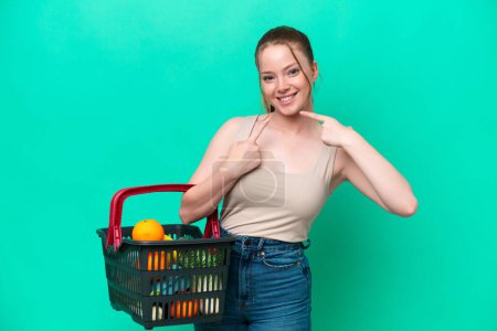 Photo for Young woman holding a shopping basket full of food isolated on green background giving a thumbs up gesture - Royalty Free Image