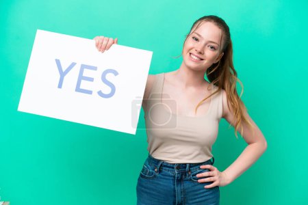 Junge kaukasische Frau isoliert auf grünem Hintergrund mit einem Plakat mit dem Text JA mit glücklichem Gesichtsausdruck