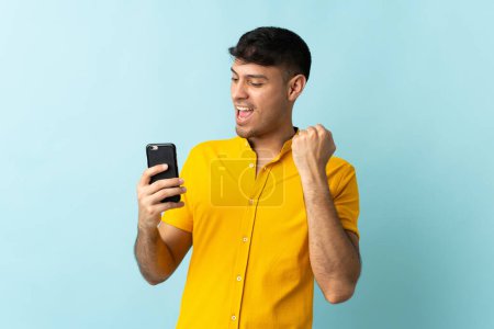 Téléchargez les photos : Jeune homme colombien utilisant téléphone portable isolé om fond bleu célébrant une victoire - en image libre de droit