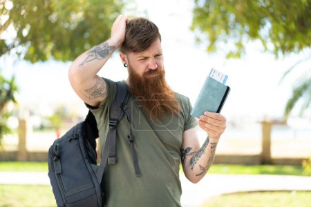 Téléchargez les photos : Rousse homme avec barbe tenant un passeport à l'extérieur ayant des doutes et avec une expression confuse visage - en image libre de droit
