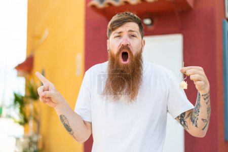 Téléchargez les photos : Homme rousse avec barbe tenant les clés de la maison à l'extérieur surpris et pointant côté - en image libre de droit