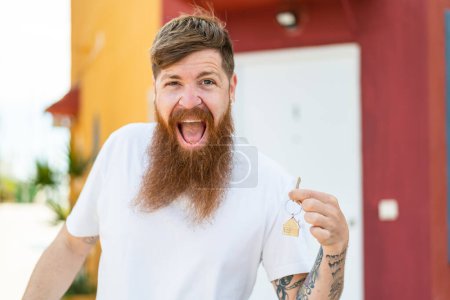 Téléchargez les photos : Homme rousse avec barbe tenant les clés de la maison à l'extérieur avec une expression faciale surprise et choquée - en image libre de droit